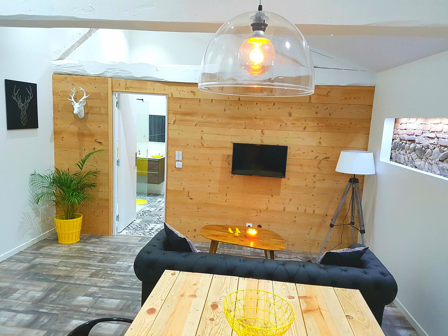 Een moderne woonkamer met een houten wand, een zwarte bank, een houten tafel, een gele plant, een lamp en een televisie.