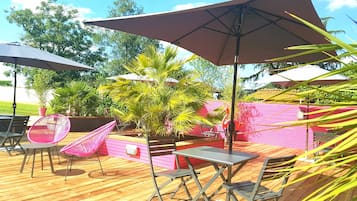 An outdoor patio with wooden flooring, metal tables, and chairs, surrounded by potted plants and umbrellas.