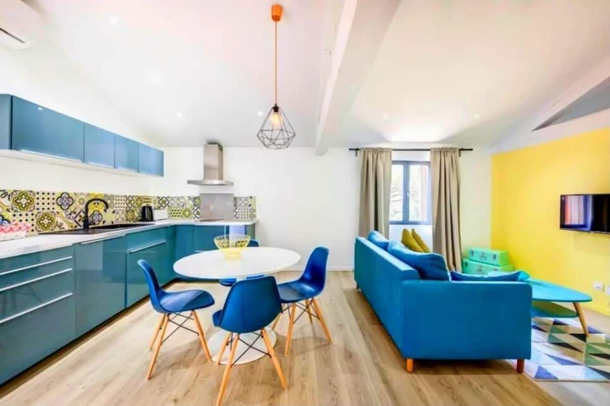 A modern kitchen with blue cabinets, a white dining table with blue chairs, and a yellow wall in the adjacent living area with a blue sofa.