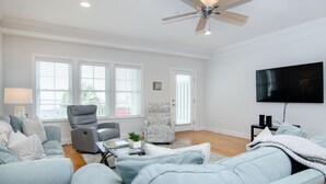 A modern living room with a grey sofa set, a recliner, a coffee table, and a flat-screen TV mounted on the wall. - 4 BR Corner Unit Oceanfront Steps to it all! (Kure Beach)