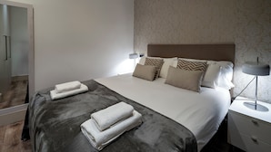 Ein ordentlich gemachtes Bett mit grauer Decke, weißen Handtüchern, einer Nachttischlampe und gemusterten Kissen.