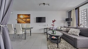 Un salon moderne avec un canapé gris, une table à manger avec des chaises, un téléviseur à écran plat fixé au mur et une plante décorative posée sur une table d’appoint.