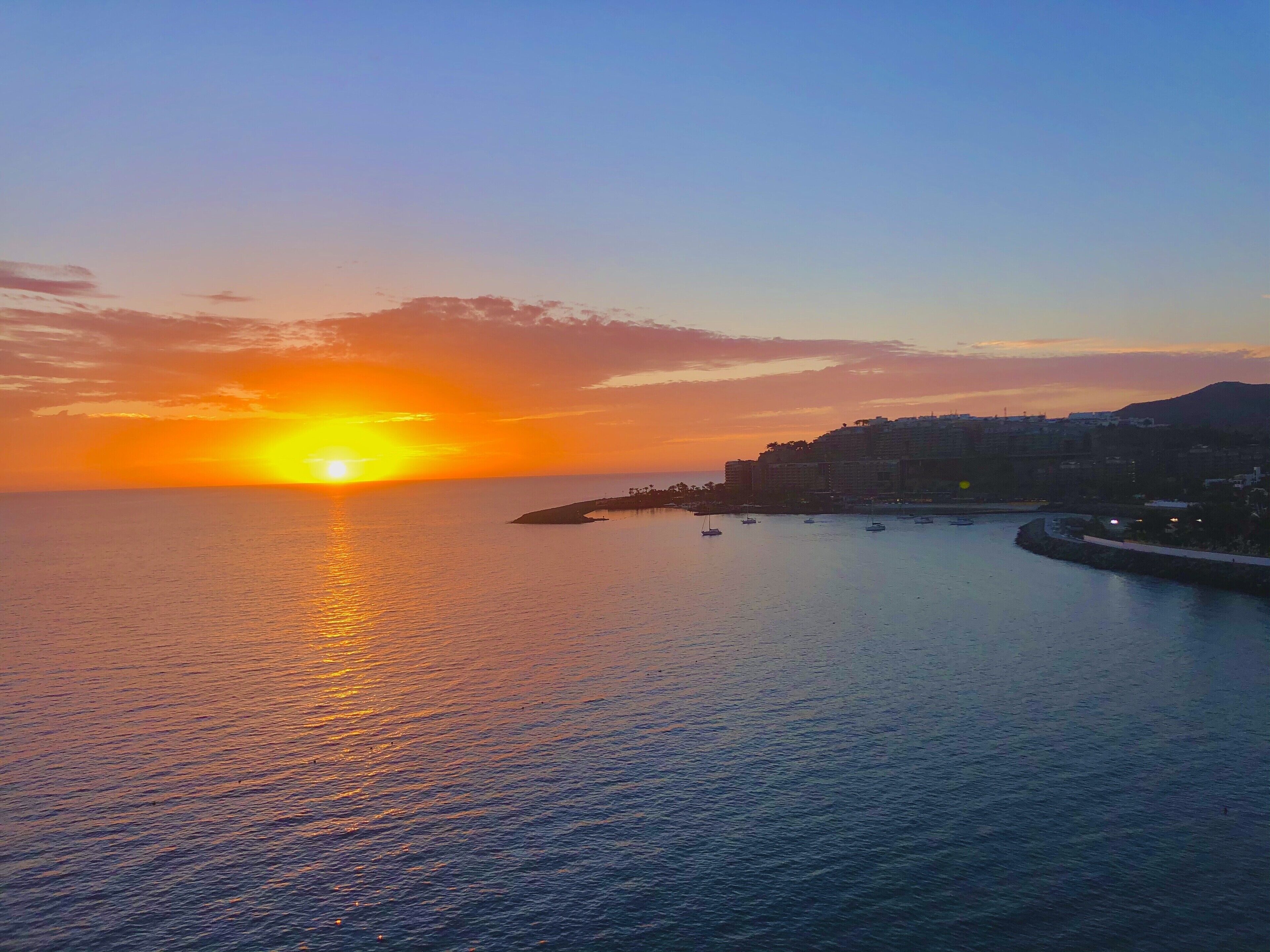 A coastal sunset with calm waters reflecting the sky, a peninsula with buildings, and a clear horizon.