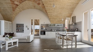 Un vaste espace de vie doté d’un haut plafond en arc de cercle, d’une table à manger avec des chaises, et d’une cuisine avec des meubles blancs et une grande fenêtre.