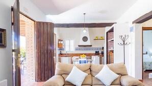 Sala de estar con un sofá color beige, una mesa de centro y vistas a una cocina con pared de ladrillo y una lámpara colgante.
