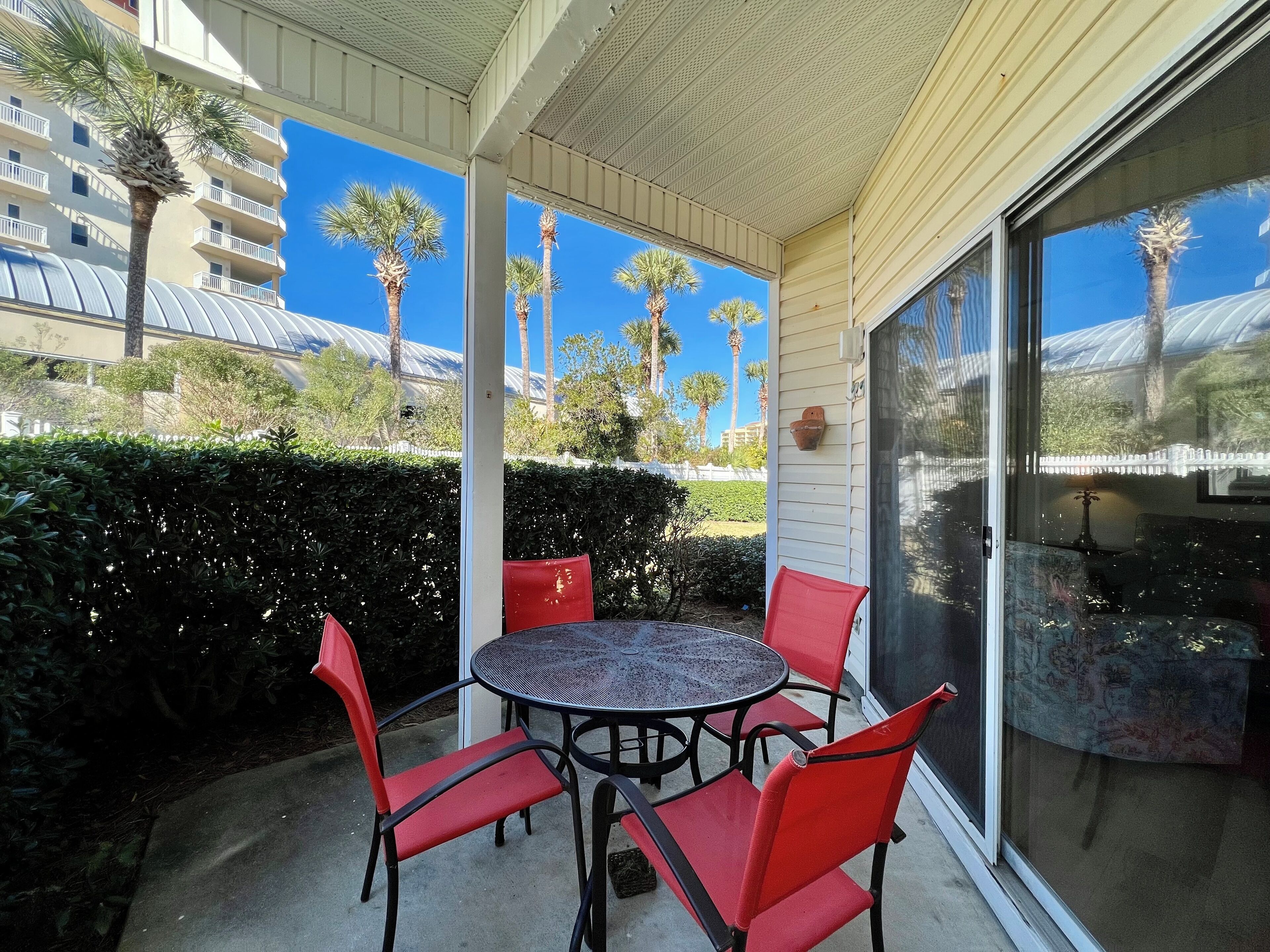 A covered outdoor patio with a round table and four red chairs, overlooking a residential area with palm trees and a white fence.