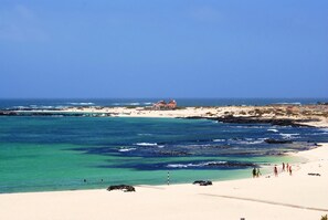 Une plage aux eaux turquoise et limpides, un rivage sablonneux et, au loin, un bâtiment coiffé d’un toit rouge.