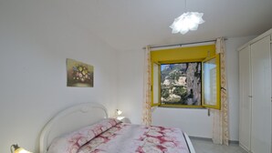 Un dormitorio con cama, lámpara de noche, ventana con marco amarillo y vista a un paisaje montañoso.