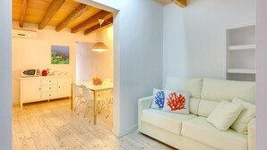 Un espace de vie moderne avec un plancher en bois, un canapé blanc agrémenté de coussins à motifs corail, un coin repas avec une table et des chaises blanches, et une cuisine équipée d’un four à micro-ondes et d’une bouilloire rouge.