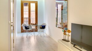 Un coin repas avec une table en verre et deux chaises blanches, un téléviseur posé sur un meuble et un tableau accroché au mur.