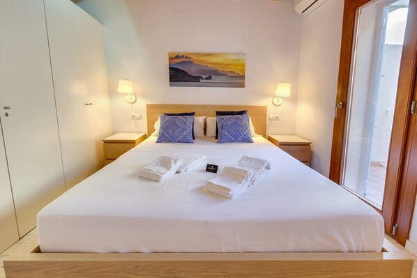 Cama bien hecha con edredón blanco, dos almohadas estampadas y toallas dobladas sobre un cabecero de madera.