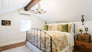 A bedroom with a bed, a chandelier, a window, a wall-mounted air conditioner, and a wooden ceiling beam.