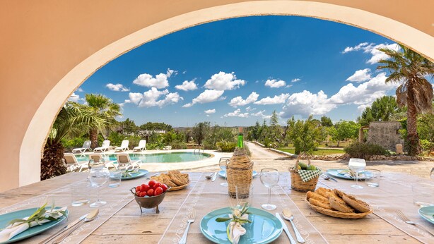 Ein Essbereich im Freien mit Blick auf einen Pool und Palmen, ausgestattet mit Tellern, Besteck und einem Brotkorb.