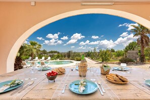 Ein Essbereich im Freien mit Blick auf einen Pool und Palmen, ausgestattet mit Tellern, Besteck und einem Brotkorb.