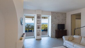 Un salon avec un canapé blanc, un téléviseur posé sur un meuble en bois et une vue sur la mer par une porte vitrée.