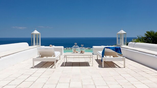 Une terrasse avec des meubles blancs, une table agr émentée d’objets décoratifs et une vue sur la mer.