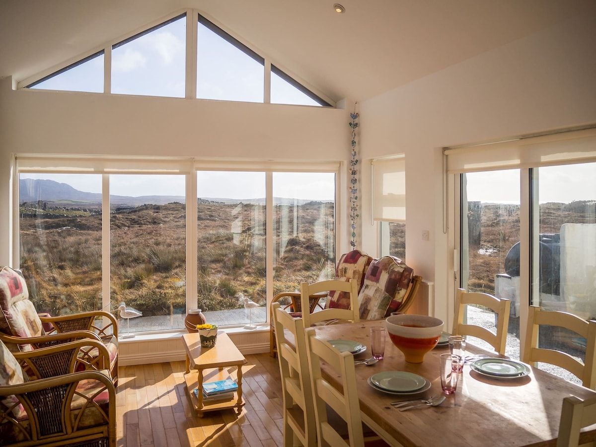 A room with a high ceiling, large windows, a dining table with chairs, and a view of a natural landscape.