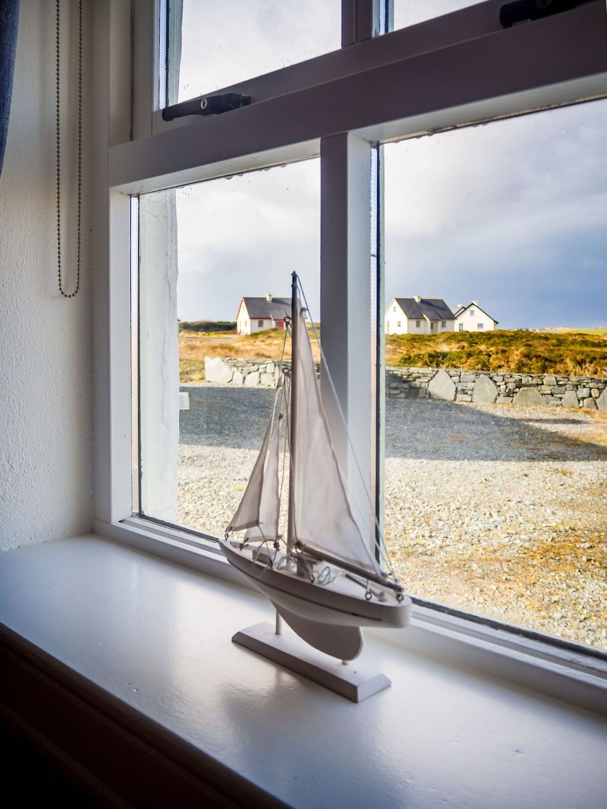 Een modelzeilbootje op een vensterbank, met uitzicht op huizen en een stenen muur.