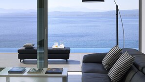 A modern living room with a black sofa, a glass coffee table, and a black floor lamp. A large window offers a view of the sea and distant mountains.