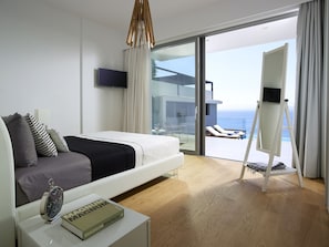 Quarto de hotel moderno com cama, mesa de cabeceira com um livro, uma janela grande com vista para o mar e varanda com cadeira e mesa.