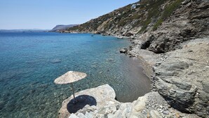 A rocky coastal area with clear blue water, a beach, and a stone structure with an umbrella.