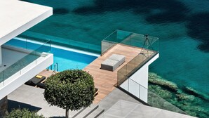 A modern building with a glass balcony overlooking a clear blue pool and the sea.
