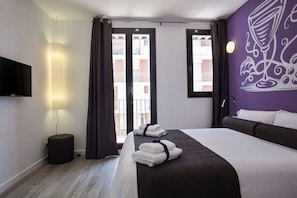 A hotel room with a bed, a TV, a lamp, and a view of buildings through the window.