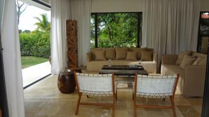 Ein modernes Wohnzimmer mit einem beigen Sofa, zwei weißen Korbstühlen, einem Couchtisch und einem großen Fenster mit Blick auf Grünflächen.