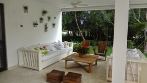 A white sofa with pillows, a wooden coffee table, and a small plant. There are two wooden stools and a white table with a chair.