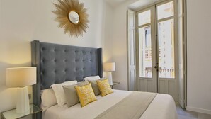 Ein Schlafzimmer mit einem gepolsterten Kopfende, zwei Nachttischlampen, einem großen Spiegel und einem Fenster mit Blick auf Gebäude.