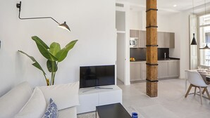 Ein modernes Wohnzimmer mit einem Flachbildfernseher, einem Couchtisch und einer Couch. Im Hintergrund ist eine Küchenzeile mit Schränken und einer Mikrowelle erkennbar.