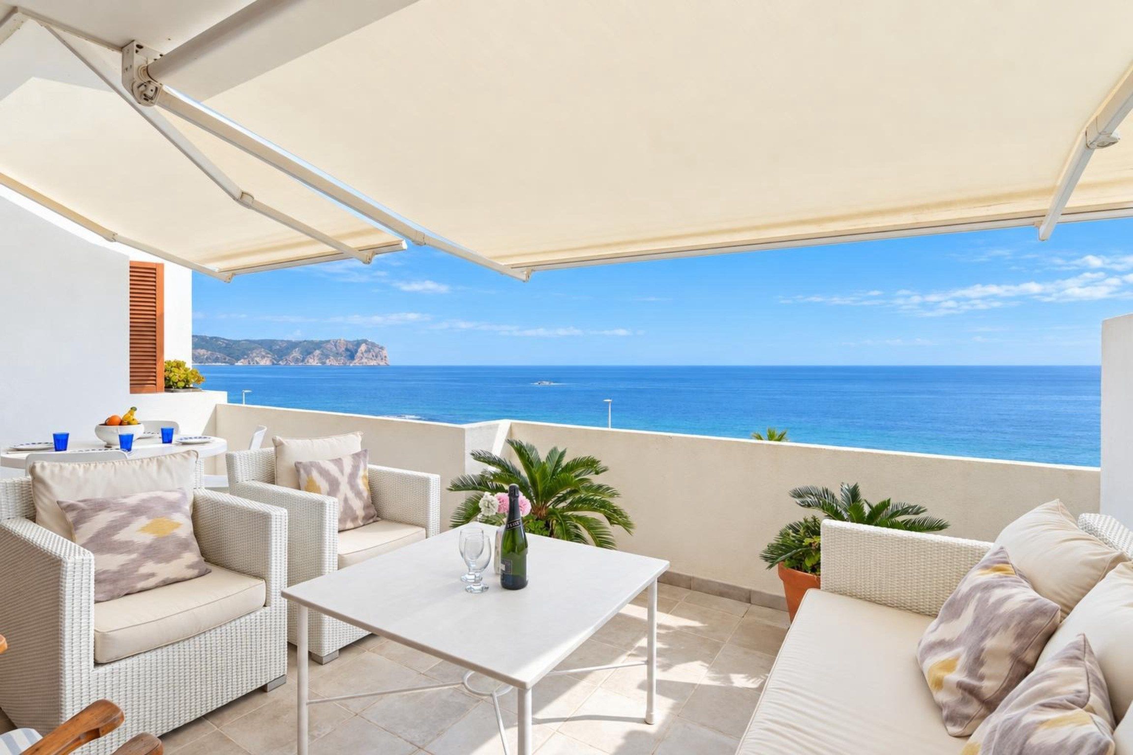 Un espacio al aire libre iluminado por el sol con vista al mar, que incluye una mesa, sillas y una planta en maceta.