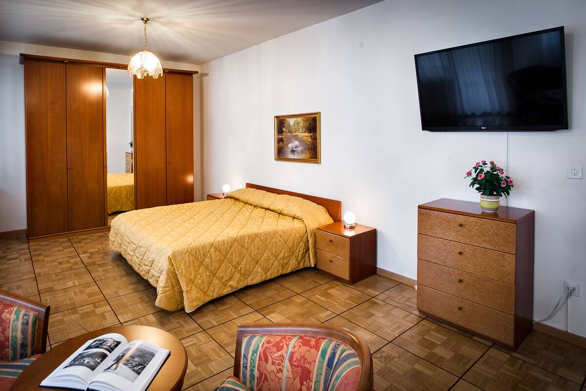 A hotel room with a bed, a TV, a dresser, a chair, and a table with a book.