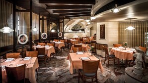 Un restaurante con varias mesas, cada una con copas de vino, platos y cubiertos. La sala tiene sillas de madera, piso de mármol y una estatua a la derecha.