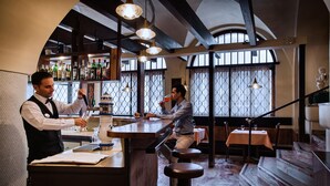 Zona de bar con un cantinero preparando bebidas, un cliente sentado en una mesa y una escalera que sube a un nivel superior.