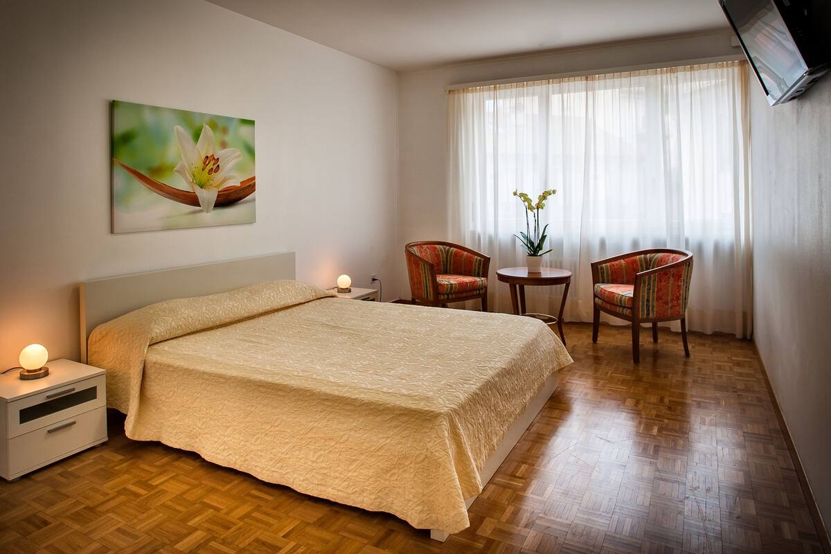A hotel room with a large bed, two chairs, a small table with a plant, a painting on the wall, and a window with curtains.