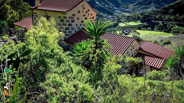 A rural house with a stone facade and red-tiled roof, surrounded by lush greenery and hills in the background. - Casa Rural Las Caldereras (Teror)