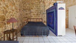 Un dormitorio con pared de piedra, una cama con manta azul, una lámpara y un refrigerador.