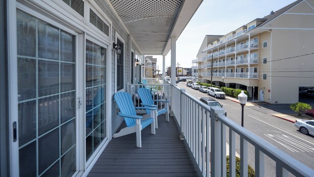 Ein Balkon mit blauen Adirondack-Stühlen, einem weißen Geländer und Blick auf eine Straße mit geparkten Autos und mehrstöckigen Gebäuden.