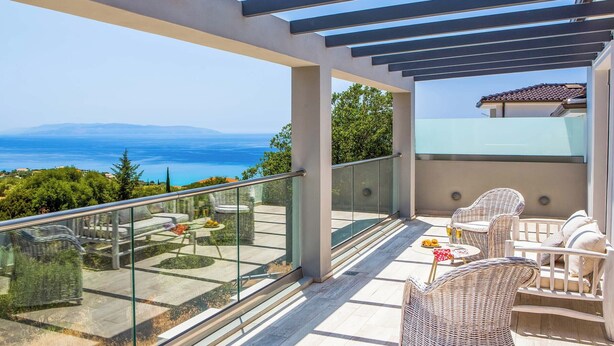 Ein Balkon mit Korbmöbeln, einer Glastrennwand und Blick auf Meer und Berge.