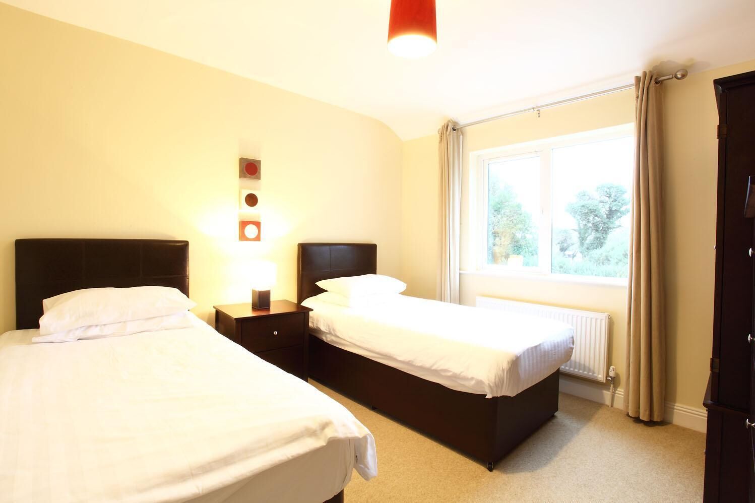 A hotel room with two beds, a window with curtains, a radiator, and a wall-mounted light fixture.