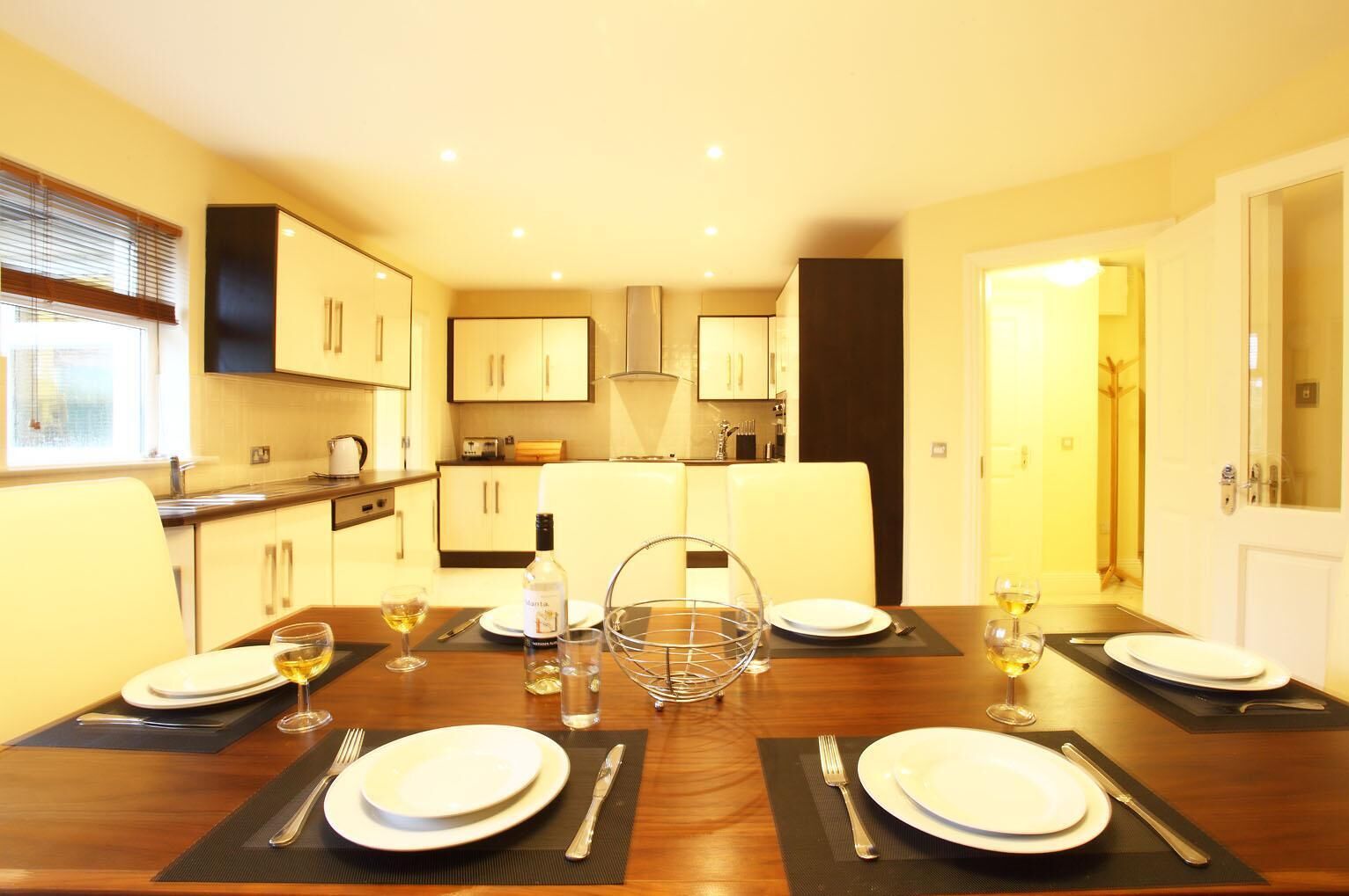 A dining area with a wooden table set for four, surrounded by white chairs. There are wine glasses, a bottle of wine, and a water pitcher on the table. The room has a modern kitchen in the background with white cabinets and a black countertop.
