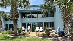 A two-story building with a light blue exterior, a covered outdoor seating area with white chairs, and palm trees. - Unit 71, Catch of The Day, 3 Bedroom Beach Condo (Panama City Beach)