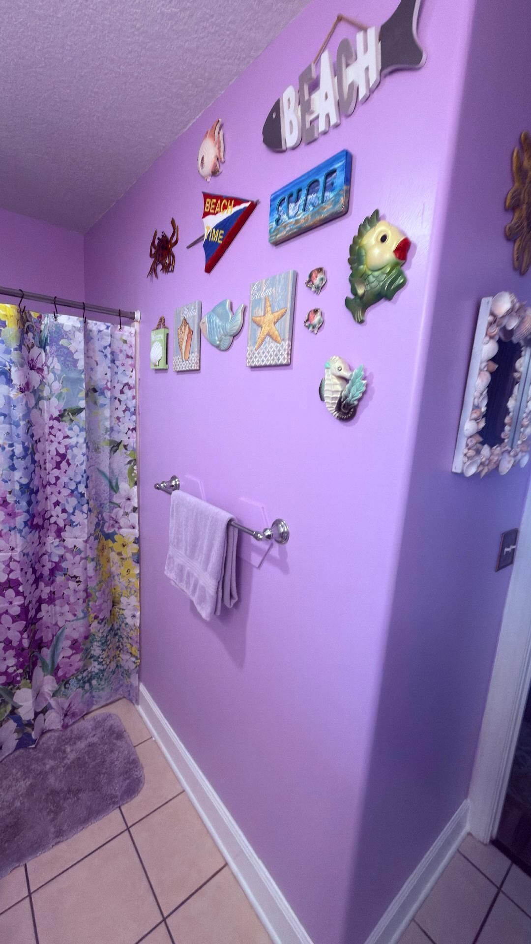 A bathroom with purple walls, a shower curtain with floral patterns, a towel rack, and various beach-themed decorations.