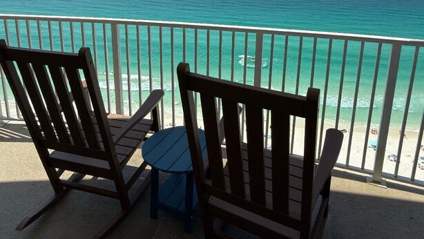 Un balcon avec deux fauteuils à bascule et une petite table, donnant sur une plage aux eaux turquoise et limpides et sous un ciel dégagé.