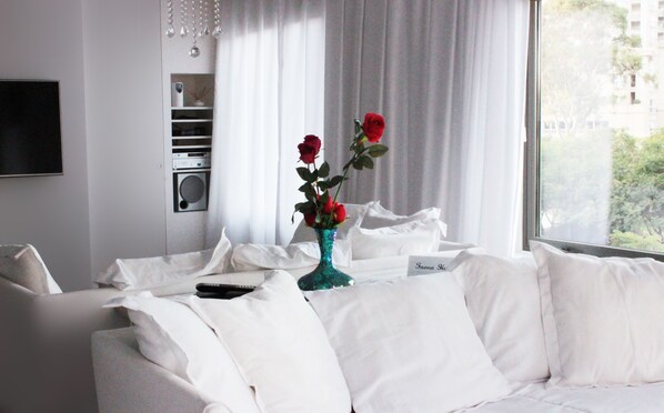 A modern hotel room with a white sofa, a teal vase with red roses, a flat-screen TV mounted on the wall, and a window with a view of buildings and trees.