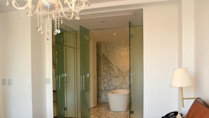 Une chambre à coucher avec un grand lit, un lustre, un dressing fermé par des parois de verre, une salle de bain en marbre et une table de chevet avec une lampe.