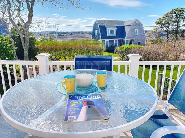 Una mesa de vidrio redonda con una revista, dos tazas amarillas y un bol sobre una terraza con barandillas blancas.