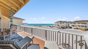 Eine Holzterrasse mit Liegestühlen und einem Tisch mit Blick auf einen Strand, im Hintergrund sind Gebäude zu sehen.