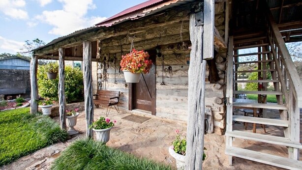 Une cabane rustique en bois, avec des fondations en pierre, une véranda ornée de pots de fleurs suspendus et un escalier en bois menant à un étage supérieur.